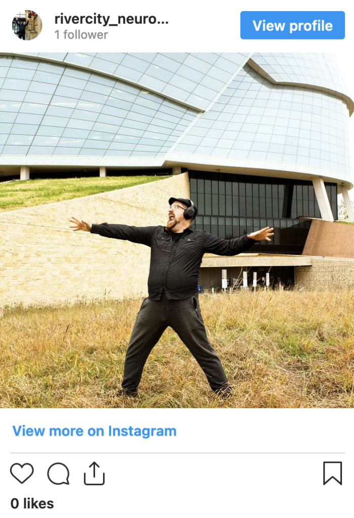 Image description of embedded Instagram post: A man dressed in all black,	 wearing a flat cap and headphones. He’s standing outside a building with lots of windows reflecting clouds in the sky. He’s stance is wide and	his arms are outstretched. The man may just be singing, or screaming?
