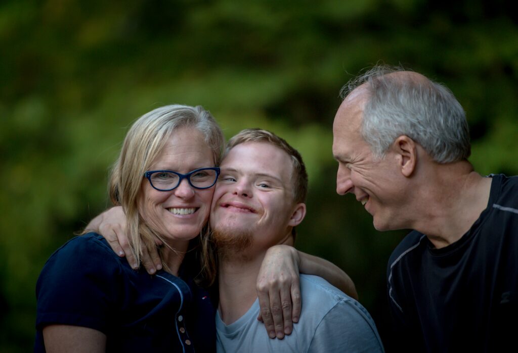 A family including a son with a visible intellectual disability, Down Syndrome. The mother is hugging the son while the father is beside them smiling and looking at the son