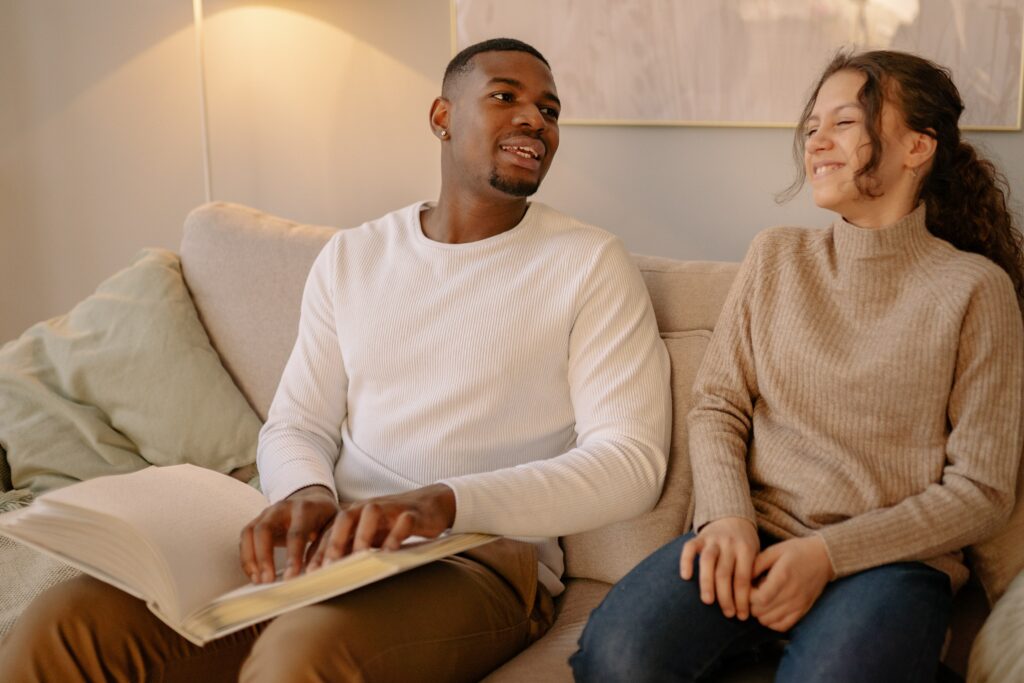 A man and woman sitting on a sofa, while the man reads a book using braille and both are smiling
