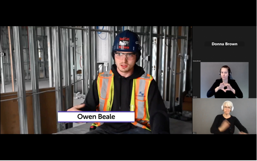Owen Beale speaks to the camera while sitting at a construction site. He is wearing a hard hat, safety glasses and a yellow and orange reflective vest. ASL and LSQ interpreters are also visible on the screen.